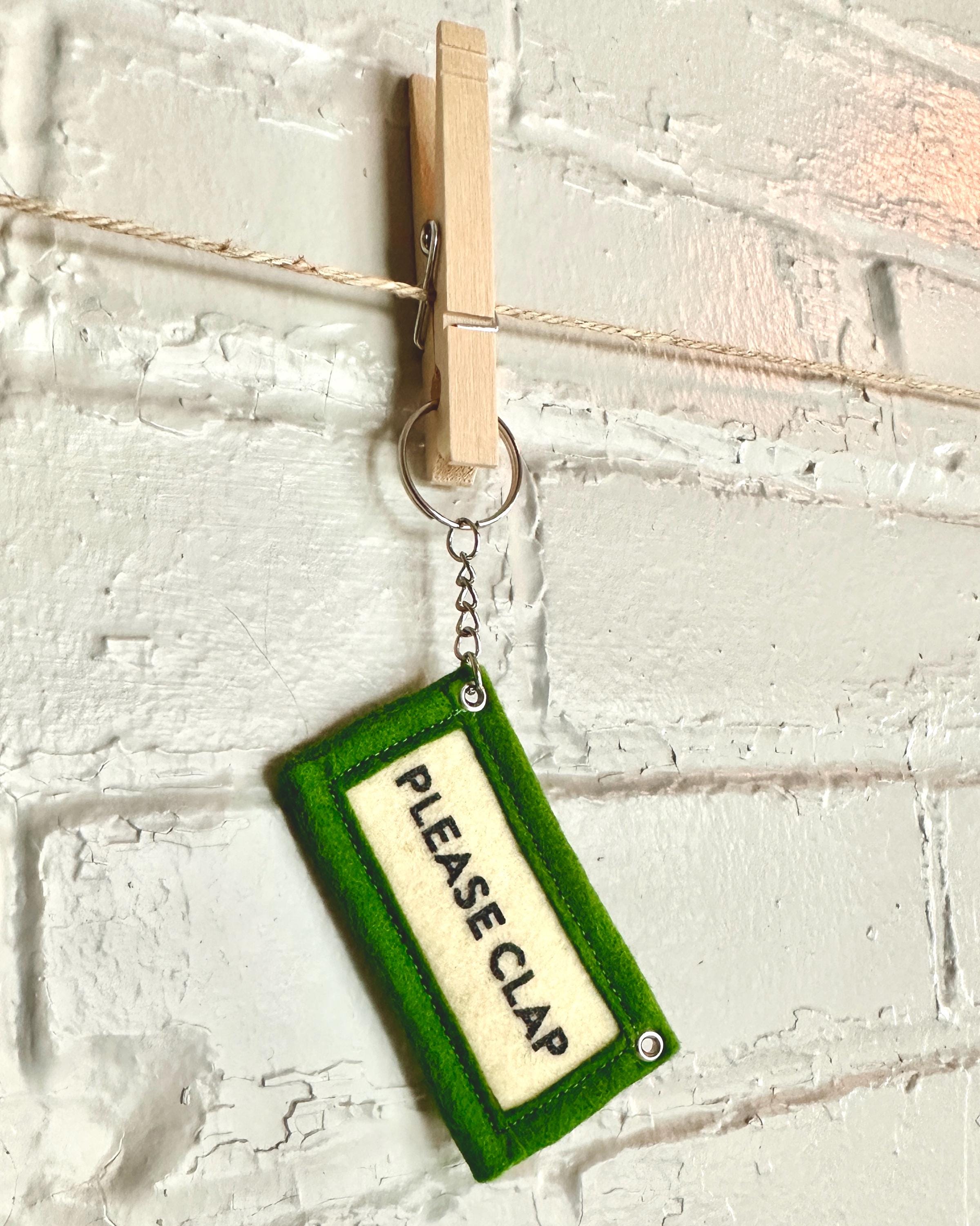 Green felt keychain with 'PLEASE CLAP' text hanging from a clothespin in front of a white brick wall