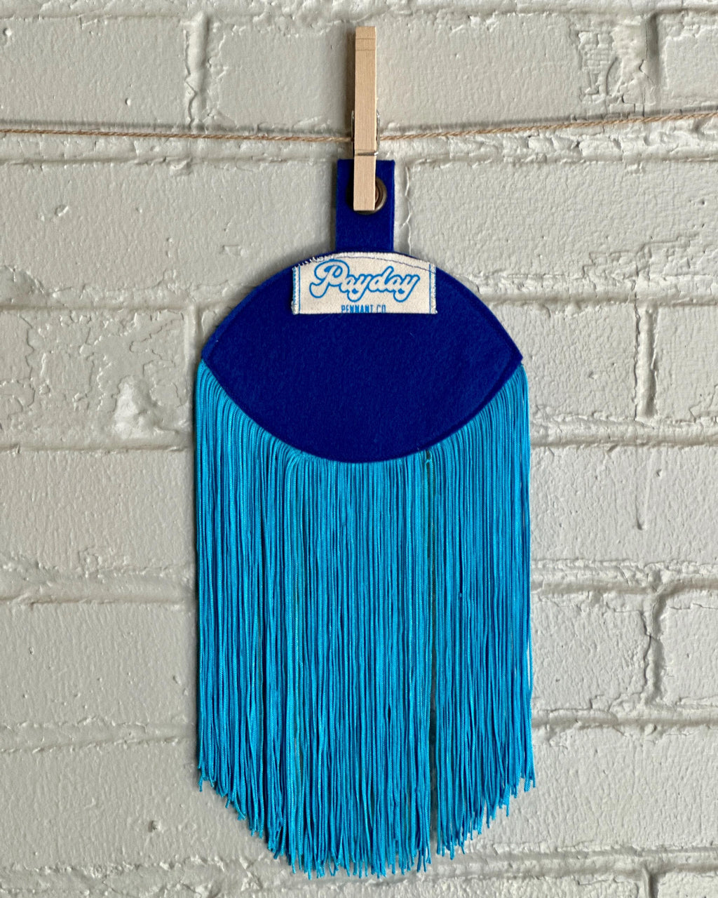 Back view of a blue felt "evil eye" with long, light blue fringe, hanging in front of a white brick wall