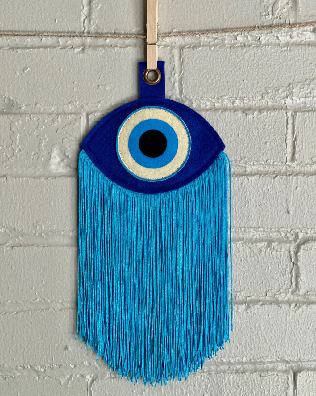 A blue felt "evil eye" with long, light blue fringe, hanging in front of a white brick wall