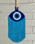A blue felt "evil eye" with long, light blue fringe, hanging in front of a white brick wall