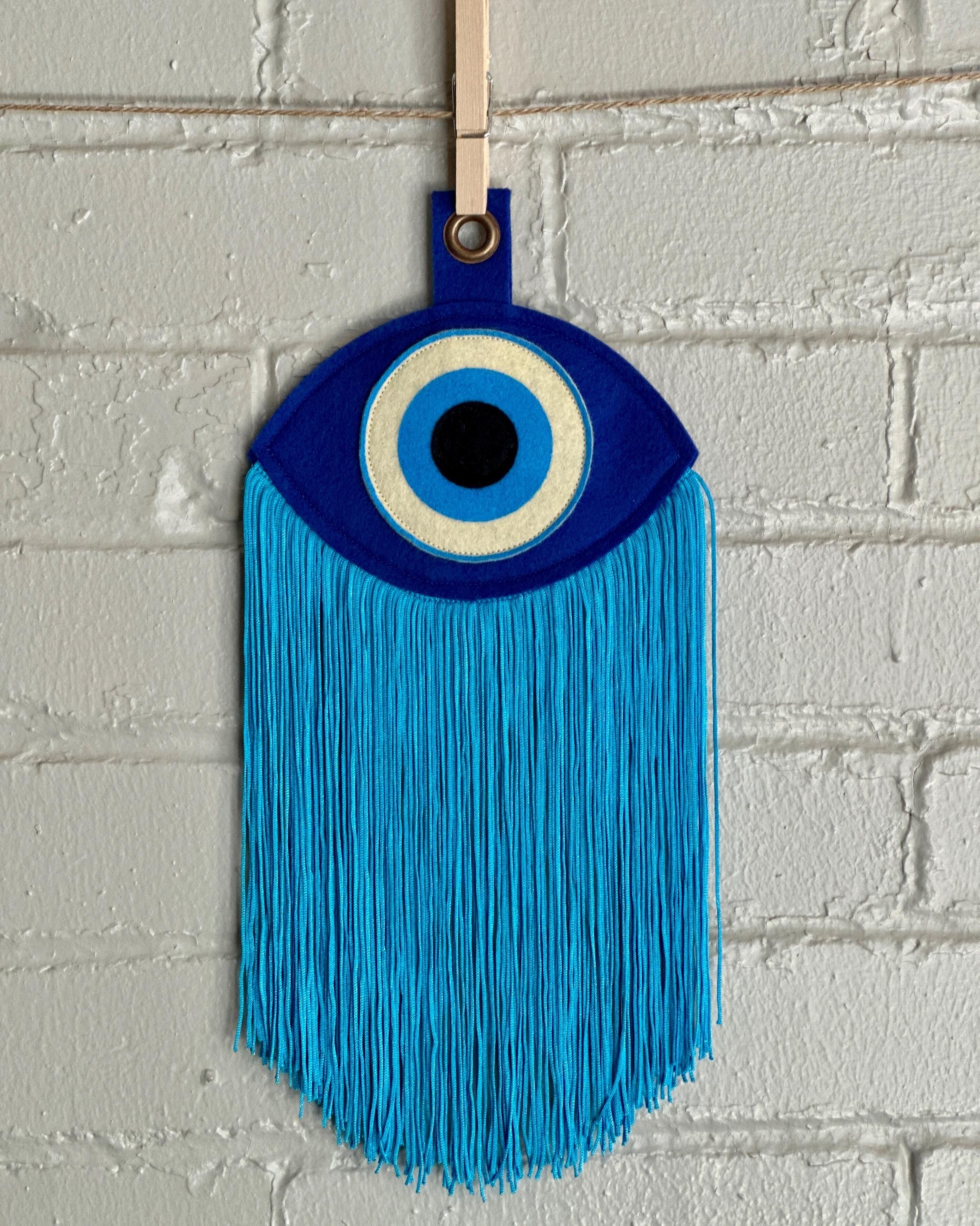 A blue felt "evil eye" with long, light blue fringe, hanging in front of a white brick wall