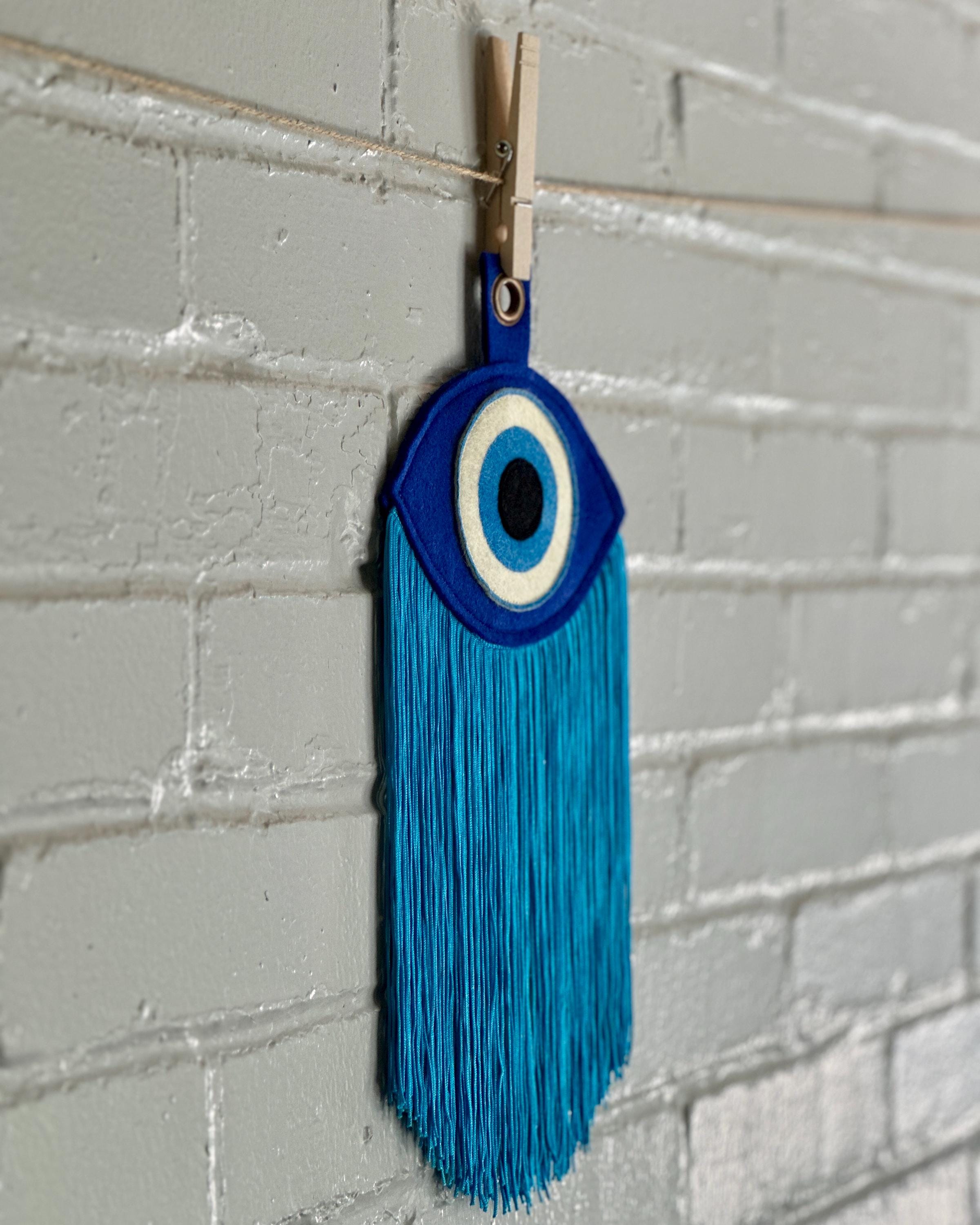 Side view of a blue felt "evil eye" with long, light blue fringe, hanging in front of a white brick wall