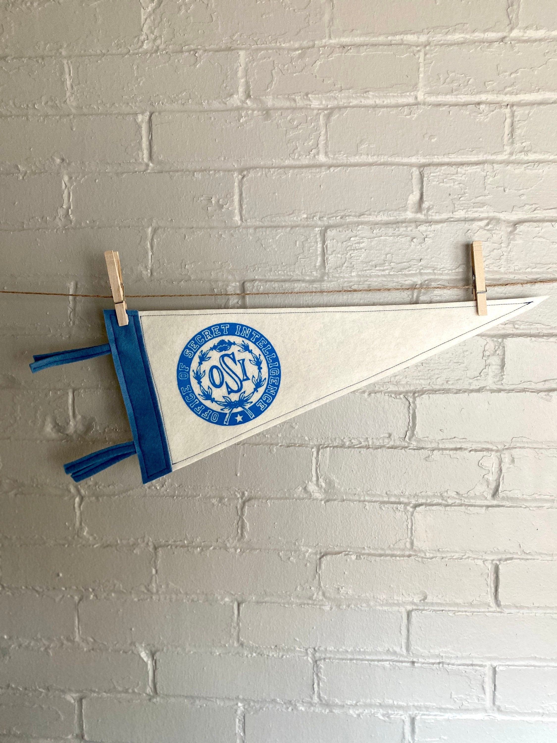 A white felt pennant with light blue trim and the OSI logo from the cartoon The Venture Bros printed in blue ink