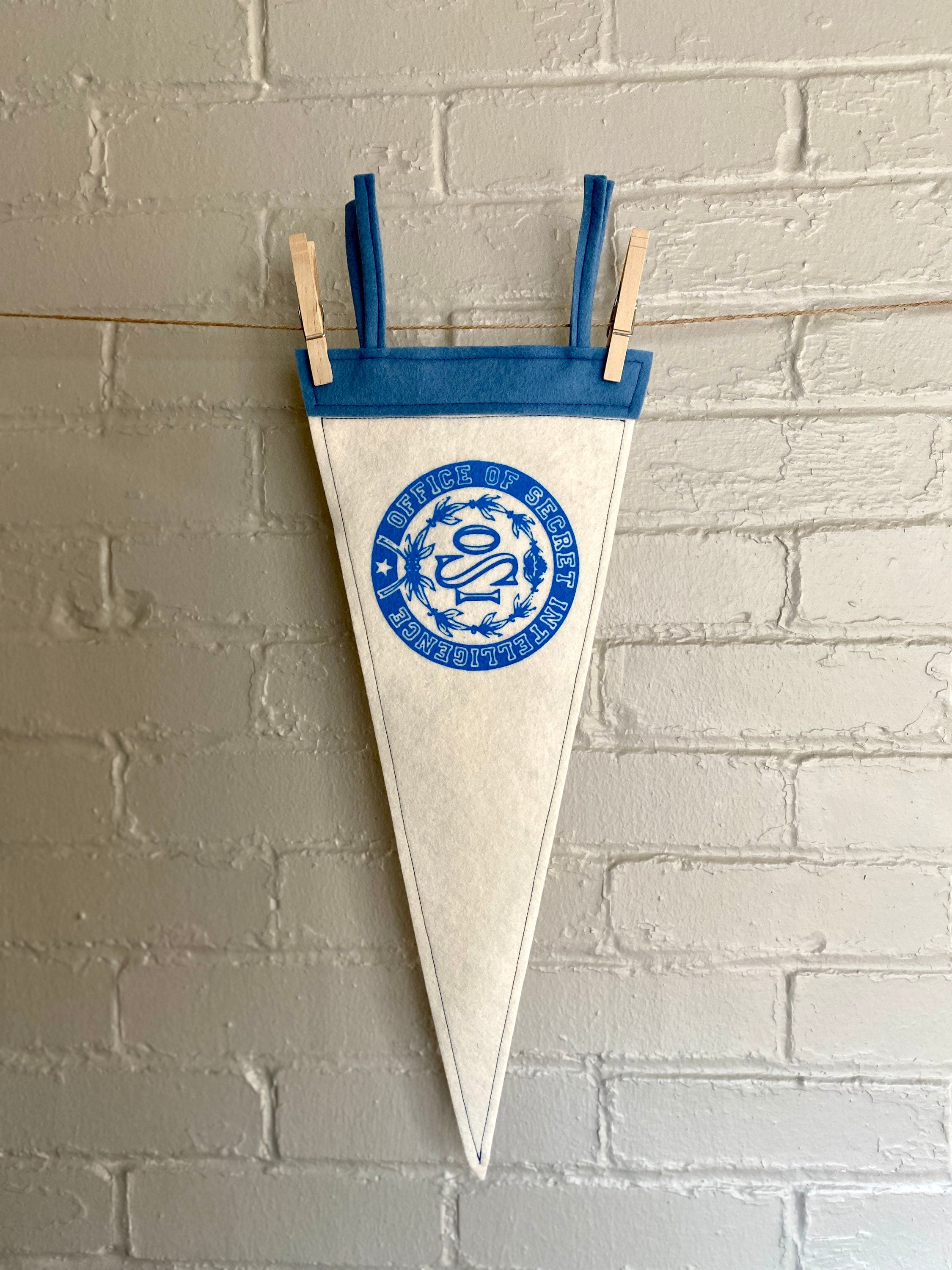 A white felt pennant with light blue trim and the OSI logo from the cartoon The Venture Bros printed in blue ink