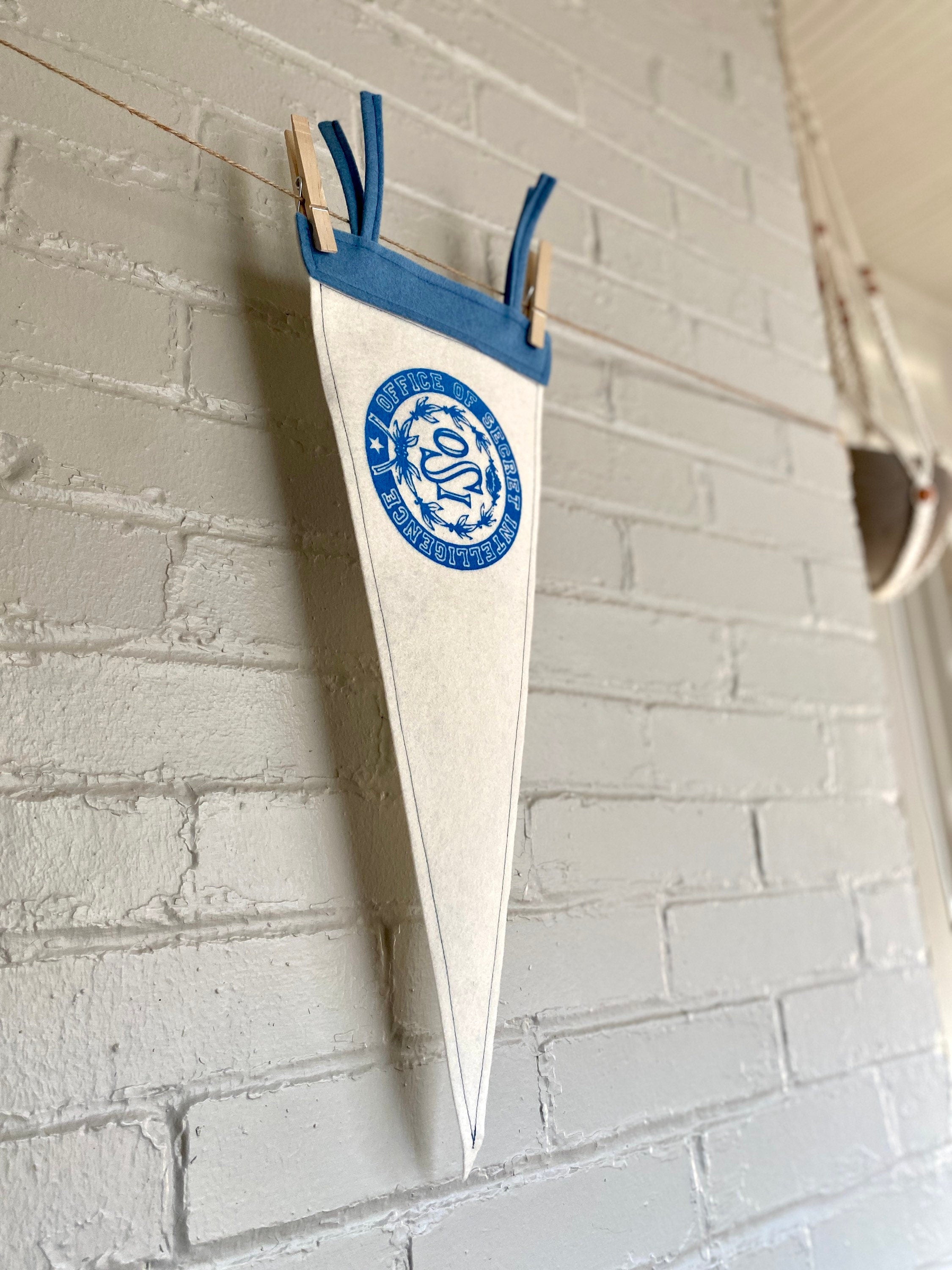 Side view of a white felt pennant with light blue trim and the OSI logo from the cartoon The Venture Bros printed in blue ink