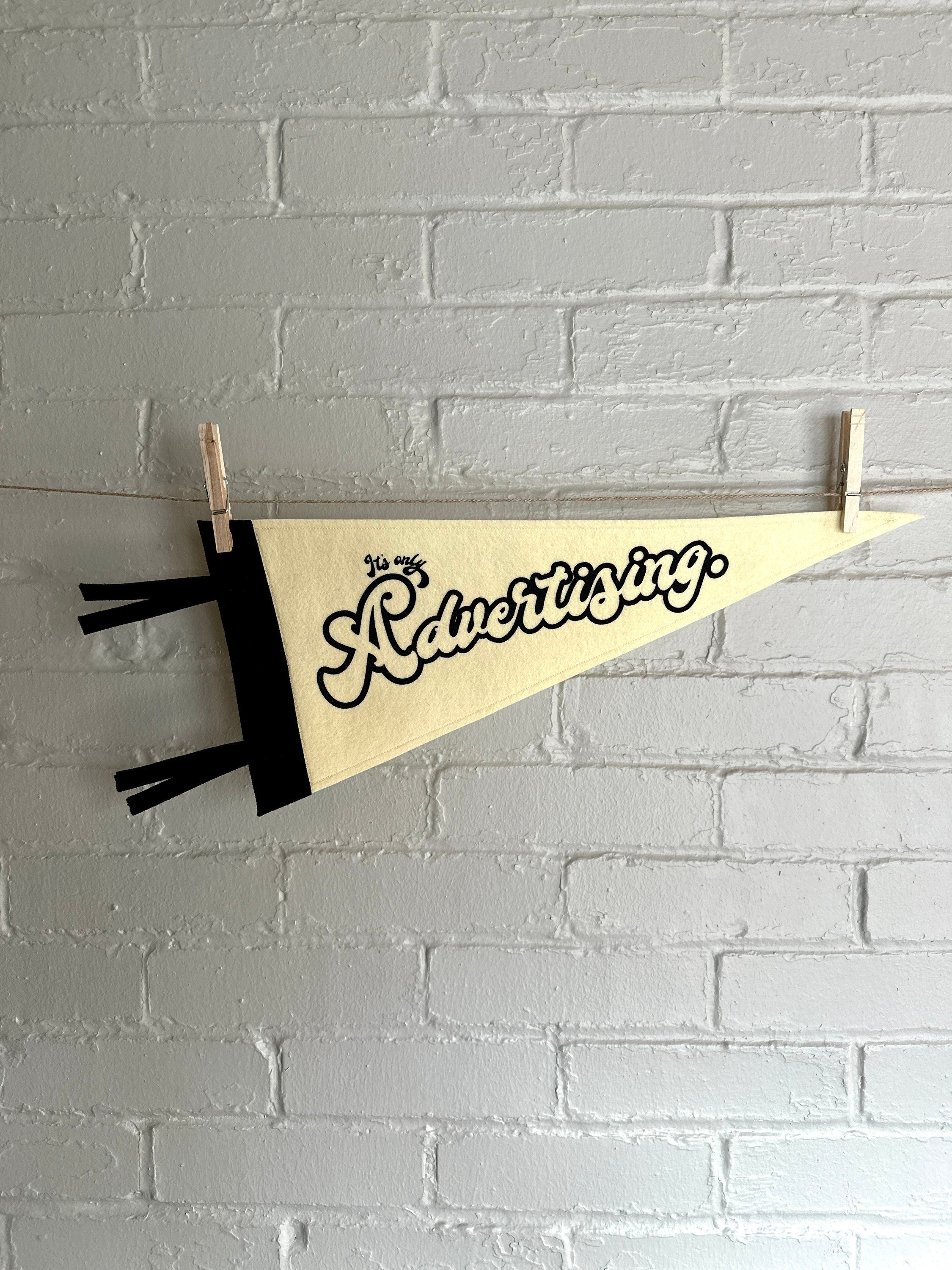 A cream felt pennant with black trim and print that reads "It's just advertising" hanging horizontally