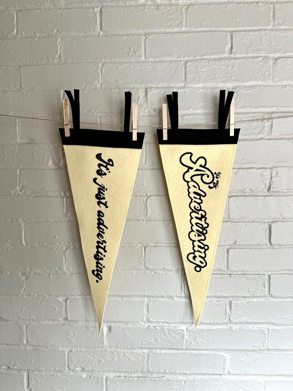 The old style "It's just advertising" pennant and new style pennant, hanging side by side on a white brick wall