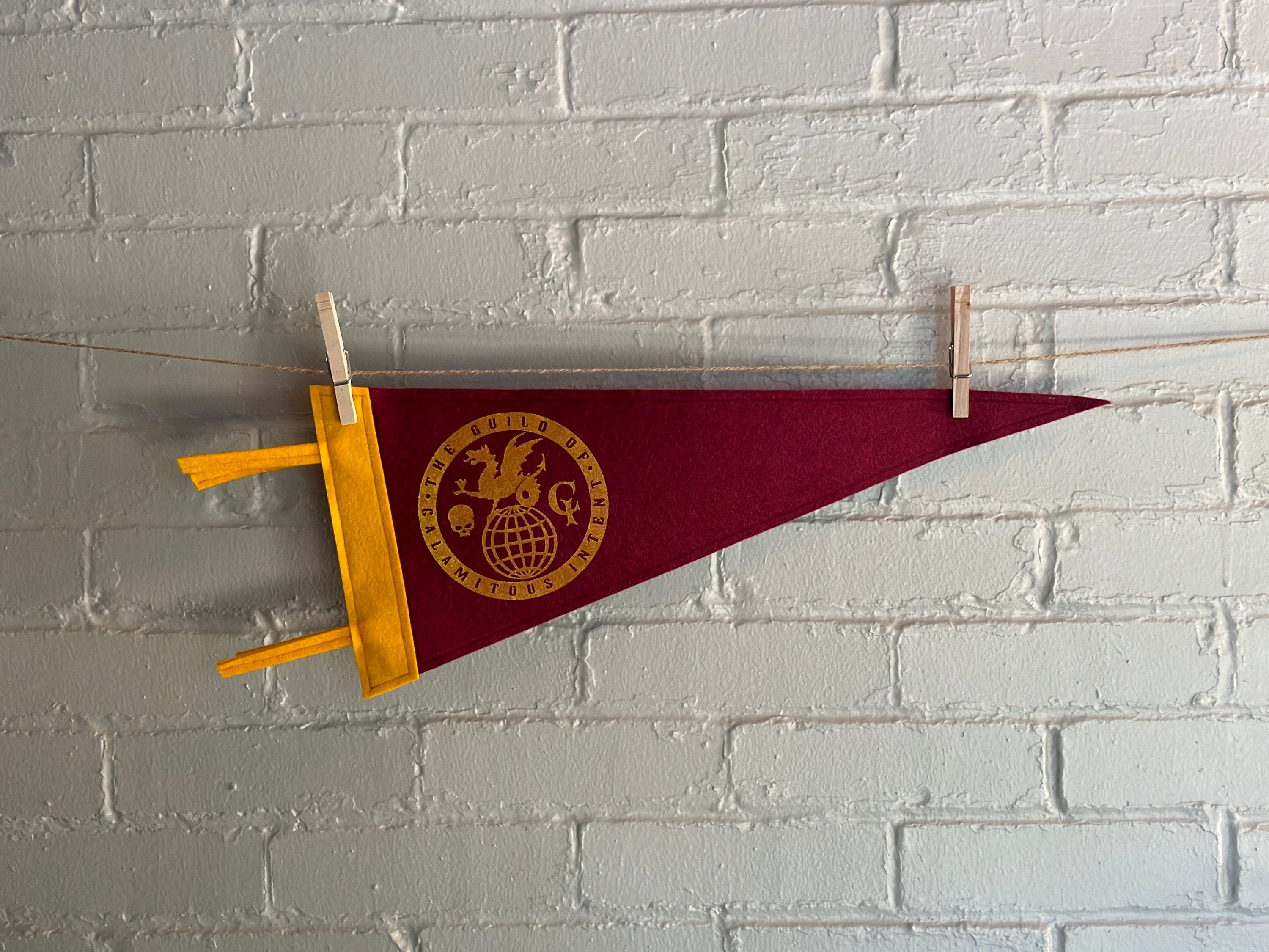 A burgundy pennant with yellow felt trim and the Guild of Calamitous Intent logo printed in yellow ink, hanging horizontally