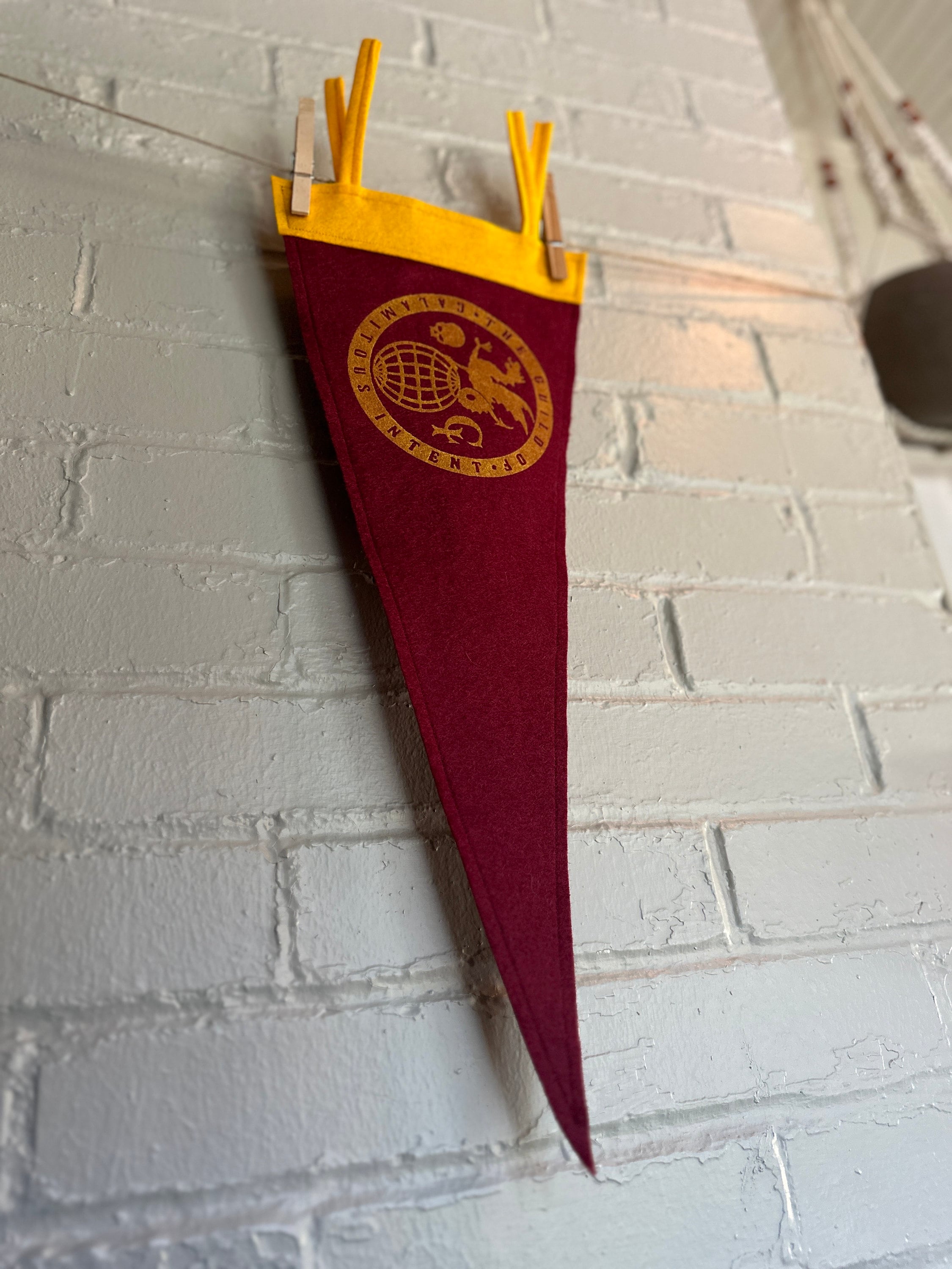 Side of a burgundy pennant with yellow felt trim and the Guild of Calamitous Intent logo printed in yellow ink
