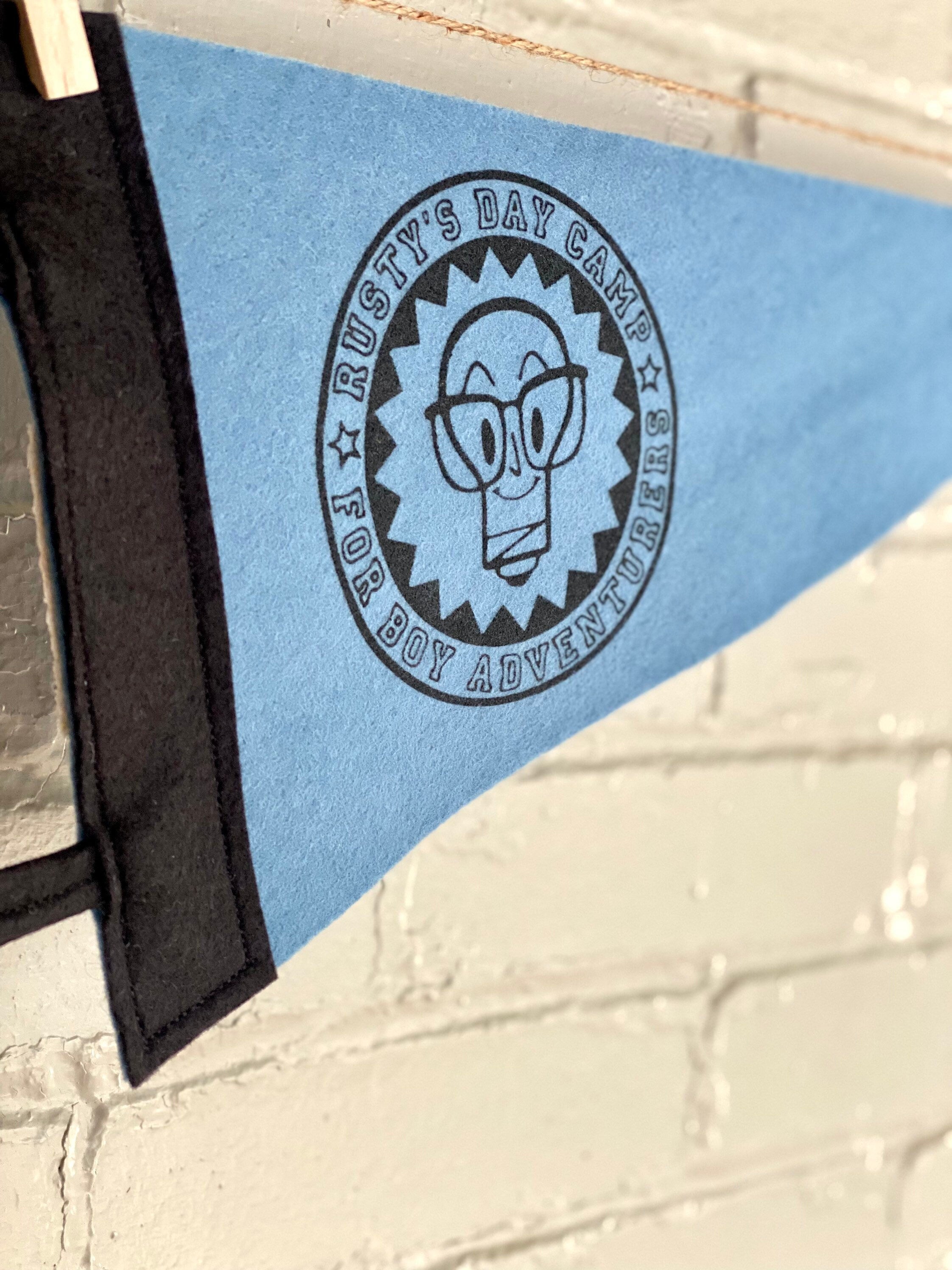 Close up of a sky blue felt pennant with black trim and a logo for "Rusty's Day Camp for Boy Adventurers" printed in black ink
