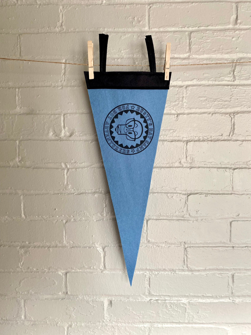 A sky blue felt pennant with black trim and a logo for "Rusty's Day Camp for Boy Adventurers" printed in black ink