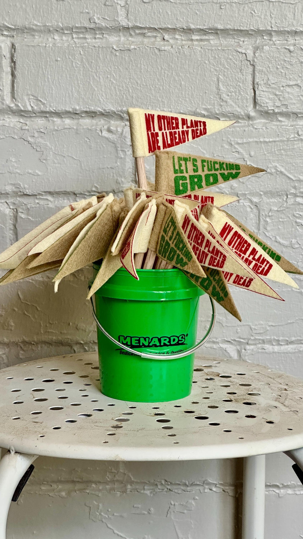 Green bucket with small pennants attached to wooden stakes, on a white stool against a gray brick wall.