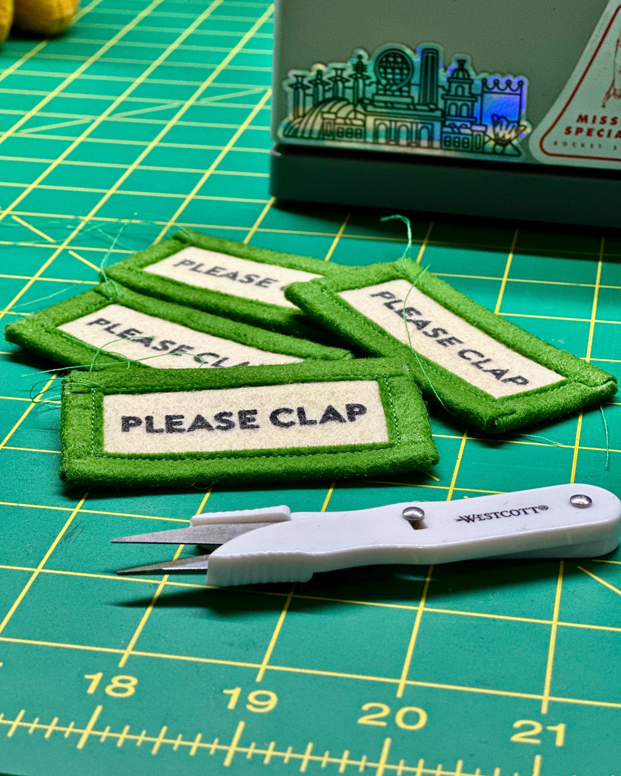 A handful of felt keychains reading "please clap" in production